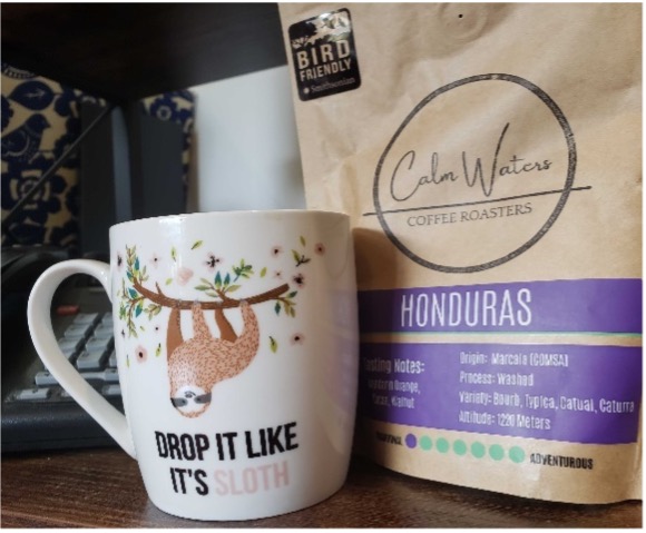 Photo of a bag of Calm Waters Roasters Bird Friendly Coffee next to a coffee mug that says "drop it like it's sloth"