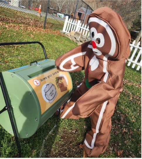 A giant gingerbread person cranks the arm of a compost tumbler