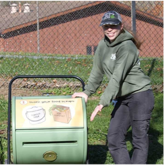 Educator Tara gesturing toward a green compost tumbler with informational and instructional decals affixed to it